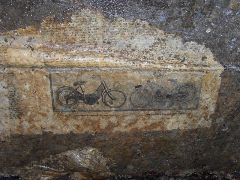 39_parallelv_archnewsp1.jpg - Newspapers on concrete arching, preserved by a thin layer of calcite. "1911 model lady's Motosacoche, with new sping fork" and "New design Moto-Reve lady's single cylinder".